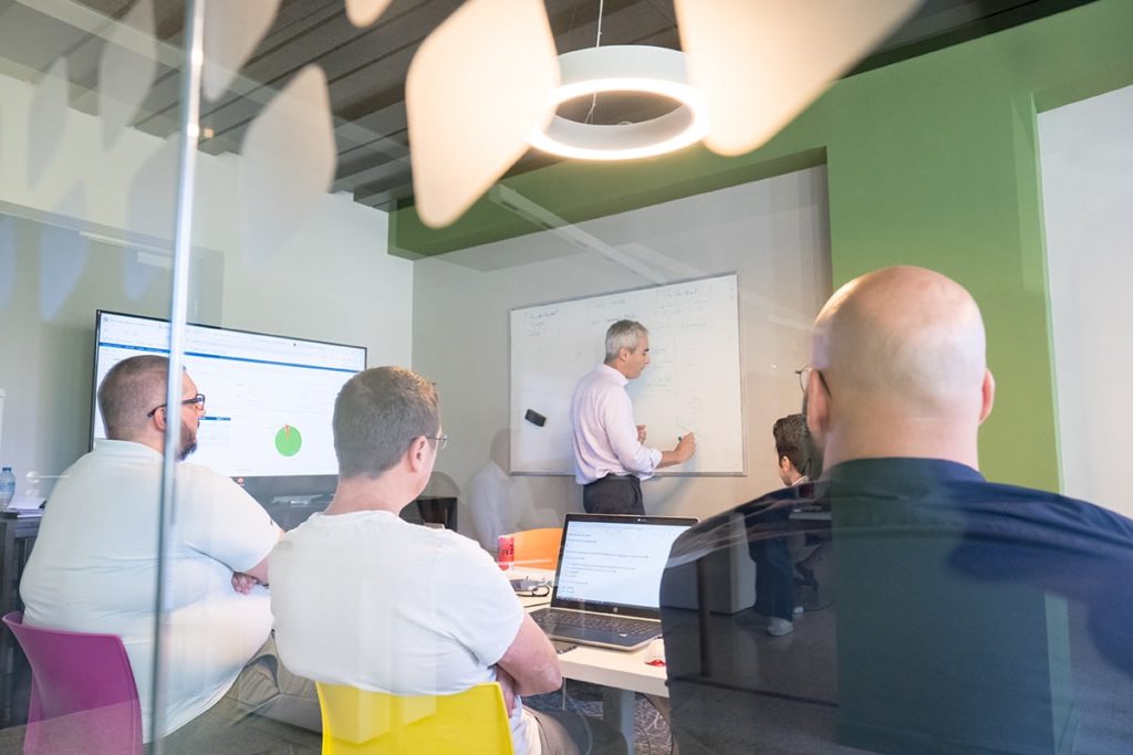 Photographie d'une formation dans les bureaux d'Arcanes. Avec un formateur réalisant un schéma sur un tableau blanc. Quatre personnes sont assis face à lui dont un client.