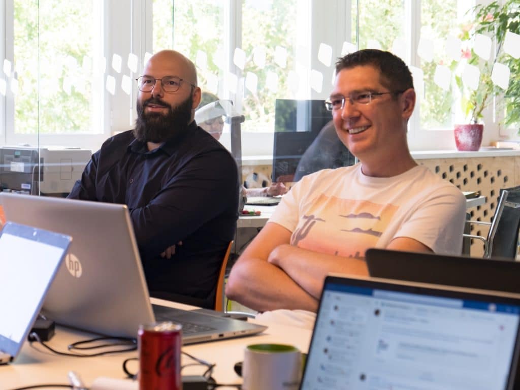 Deux personnes masculines se trouvant au bureau. Ils regardent face à eux avec un sourire. Deux ordinateurs se trouvent face à eux.