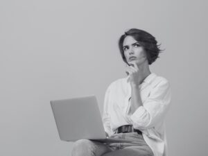 Une femme assise pensive. Elle porte une chemise blanche et un jean bleu, et elle tient un ordinateur, posé sur ses genoux.